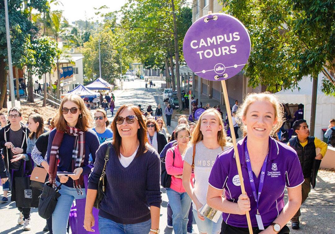 A student in a purple UQ shirt holds a "Campus tours" sign and leads a group of prospective students around St Lucia