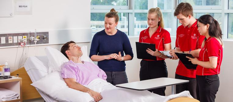 A patient lays in a hospital bed with UQ speech pathology students standing near them with clipboards