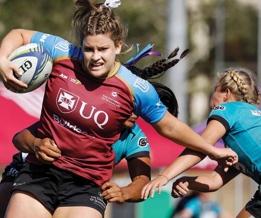 Student in blue and maroon UQ jersey holding the football and dodging a tackle in a game of women's rugby.
