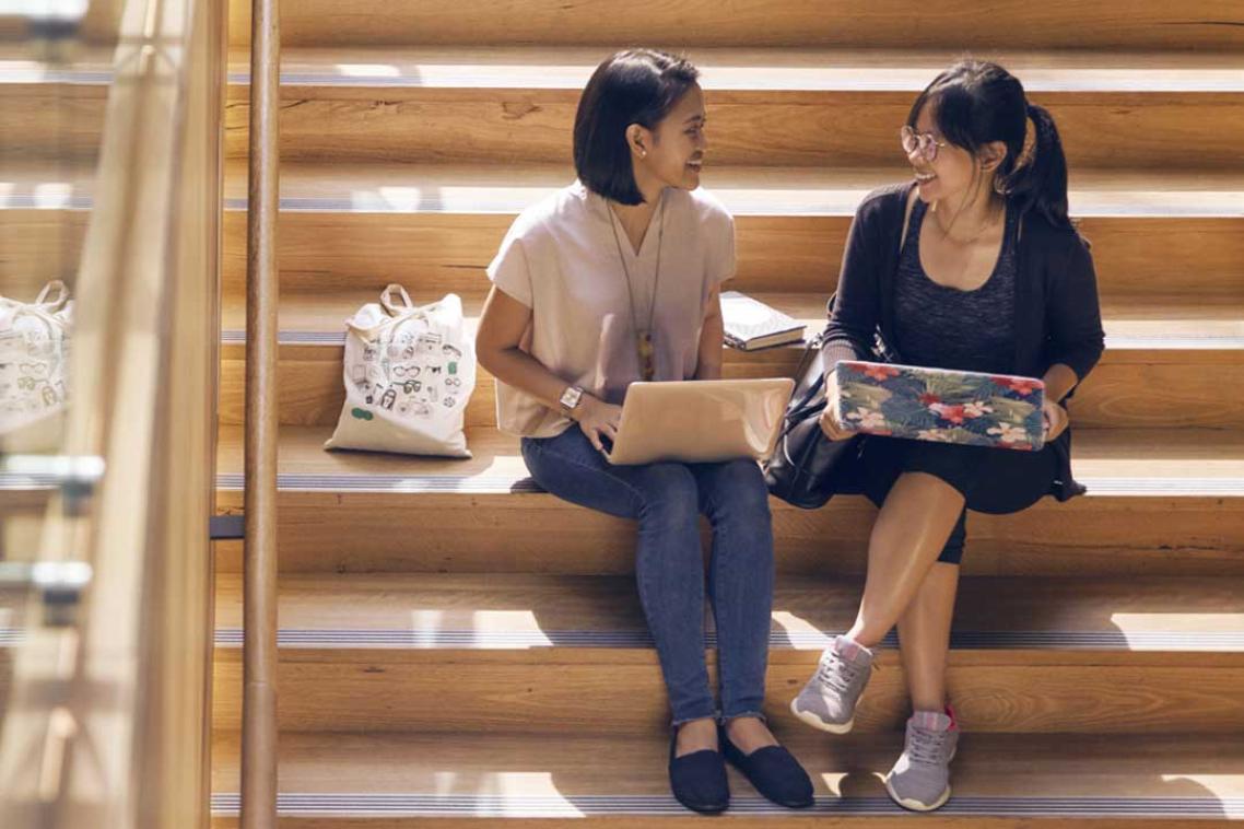 Two students working on laptops sit on a set of stairs chatting to each other
