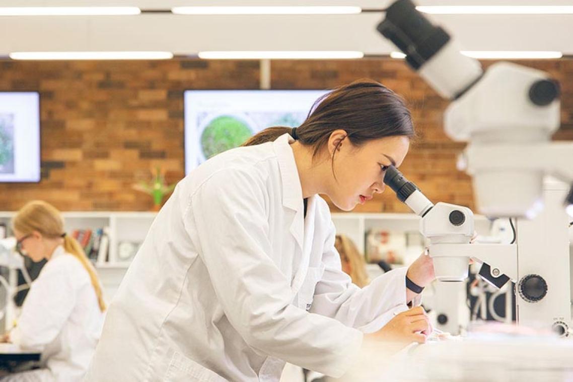 A student at Gatton campus looking through a microscope. She's wearing a white lab coat and other students work in the background.