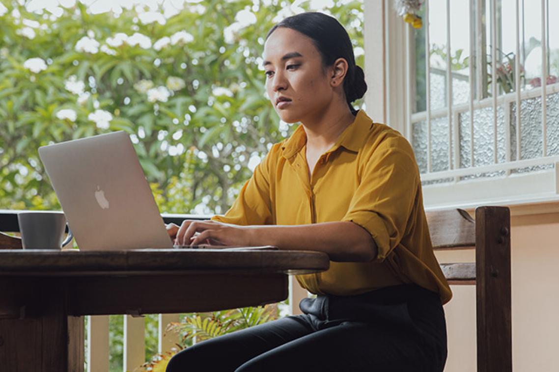 A young woman sits at a table looking seriously at her laptop screen, awaiting her university offer through QTAC.