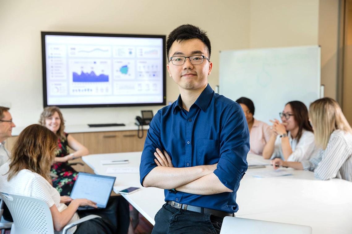 Lecturer looking directly into camera while students behind him are working